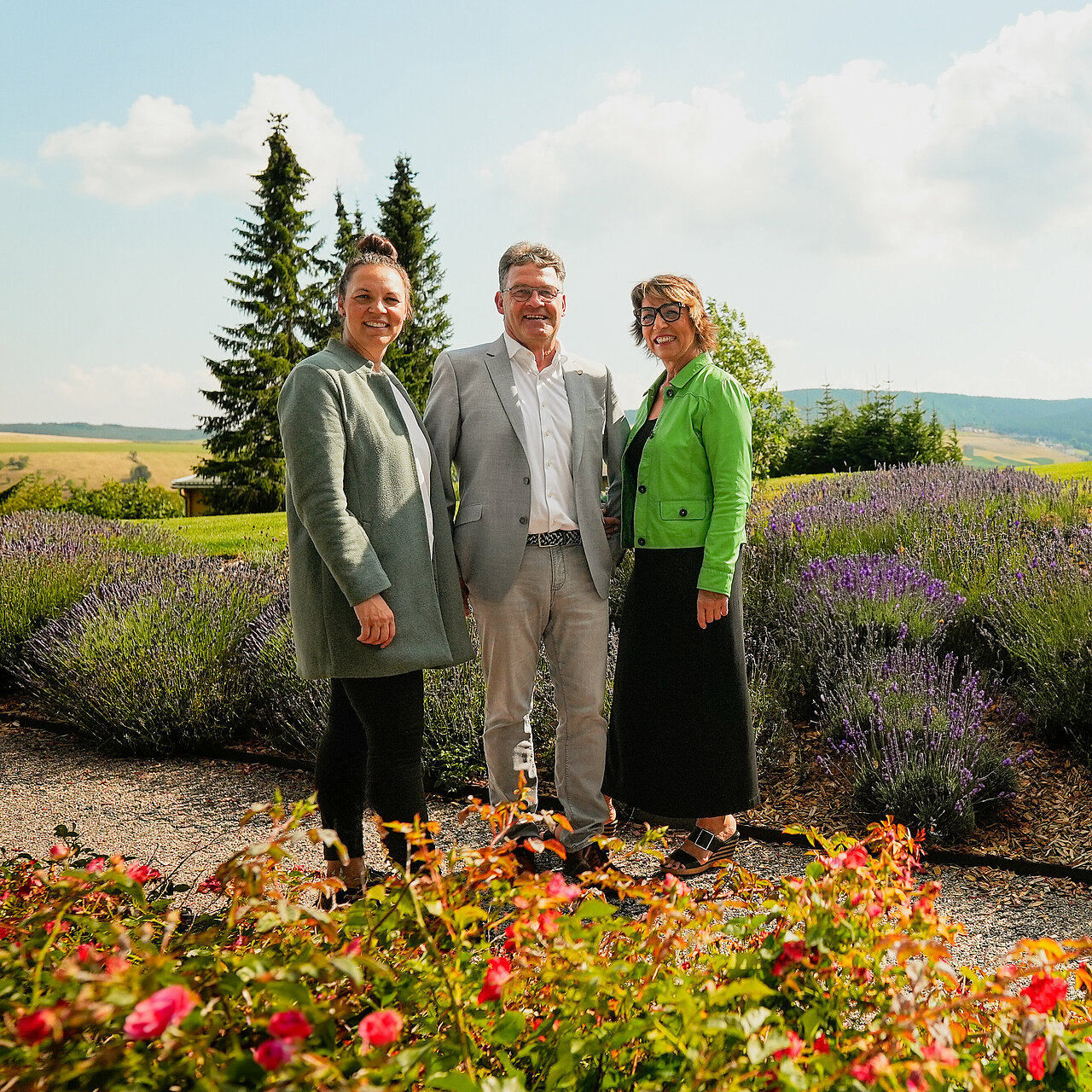 Die Gastgeberfamilie Ellinger & Duske Die Gastgeberfamilie Ellinger & Duske stehend lächelnd neben dem Spa Bereich vor dem eindrucksvollen Erzgebirgspanorama. Vor ihnen blühen zarte rote Rosen, hinter ihnen duftender Lavendel.